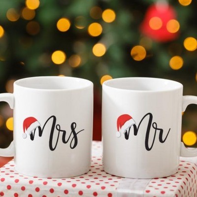 Duas canecas brancas com as palavras 'Mrs' e 'Mr' com chapéus de Natal vermelhos, sobre caixa embrulhada com bolinhas vermelhas e luzes de Natal ao fundo