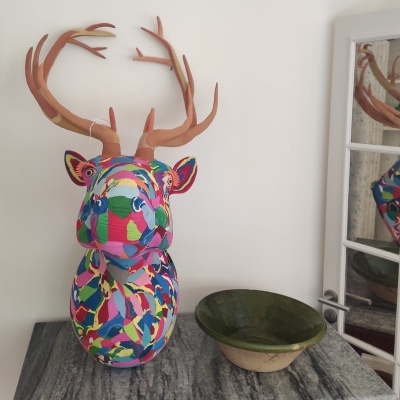 Cabeça decorativa colorida de veado com galhadas sobre mesa cinza e taça verde ao lado