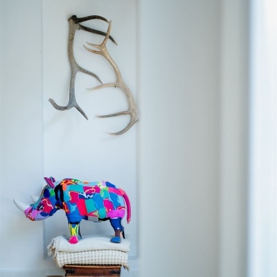 Estatueta decorativa de rinoceronte colorido sobre banco com mantas, parede branca e galhadas na parede