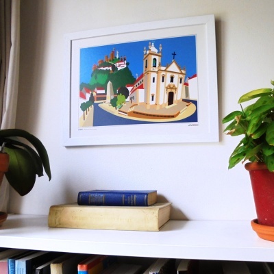 Quadro colorido de igreja e colina numa parede branca com livros e plantas em estante branca