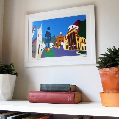 Quadro colorido de rua urbana com estátua e edifícios em moldura branca sobre estante com livros e plantas