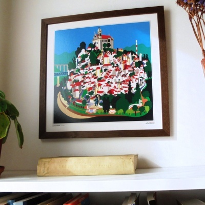 Quadro de vila colorida em moldura de madeira na parede, acima de estante branca com livro e plantas ao lado.