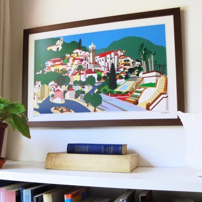 Quadro colorido de vila com moldura castanha na parede acima de estante branca com livros e planta