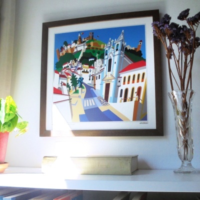 Ilustração colorida de paisagem urbana em quadro com moldura castanha sobre prateleira branca com plantas e livros