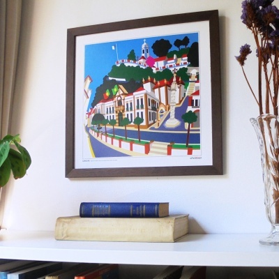 Quadro decorativo com ilustração colorida de Coruche na parede acima de prateleira branca com livros e vaso de flores secas