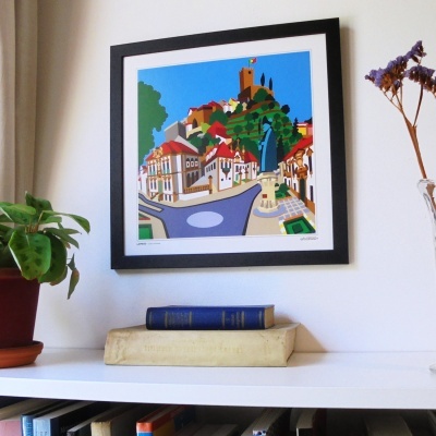 Quadro colorido com moldura preta a mostrar vila com castelo e casas, sobre prateleira branca com livros e planta