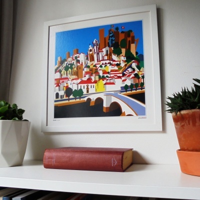 Quadro estilizado de vila com castelo sobre estante branca com livro castanho e duas plantas