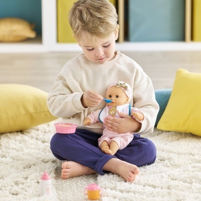 Menino a alimentar uma boneca sentados no tapete junto a acessórios de brincar