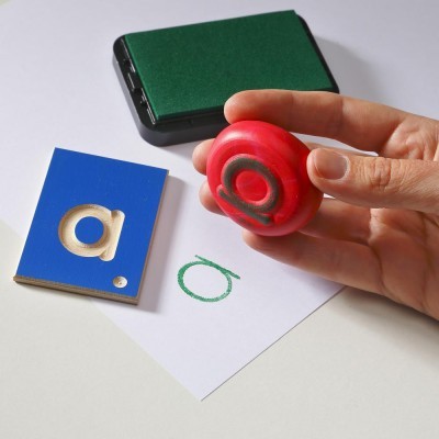 Carimbo vermelho com símbolo '@', almofada verde, bloco azul e papel branco com símbolo desenhado