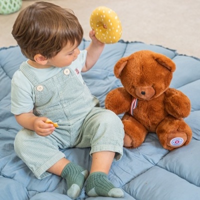 Criança com roupa verde clara segurando brinquedo amarelo junto a urso de peluche castanho num tapete azul