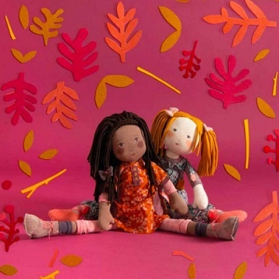 Duas bonecas de pano com vestidos coloridos sentadas em fundo rosa com folhas decorativas de feltro