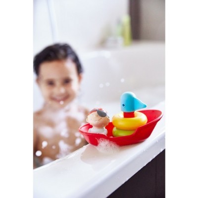 Barco de brinquedo vermelho com dois bonecos coloridos na borda da banheira branca com criança ao fundo