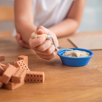 Colher adaptada com cabo de madeira e metal, taça azul com papa e blocos de construção numa mesa de madeira