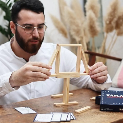 Homem monta estrutura em madeira clara com caixa KAPLA ao lado numa mesa de madeira.