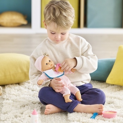 Criança a alimentar boneca de plástico com roupa rosa, sentada em tapete macio com brinquedos de alimentação.