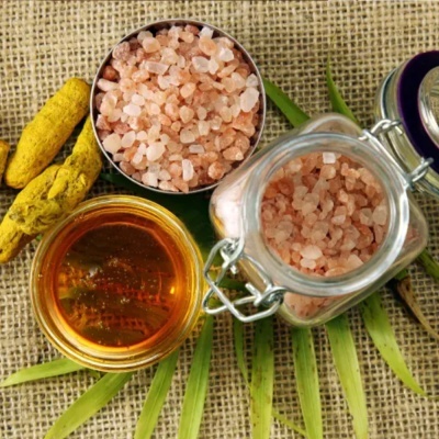 Sal grosso rosa em frascos de vidro e metal, líquido âmbar em pequeno frasco, folhas verdes sobre tecido saco