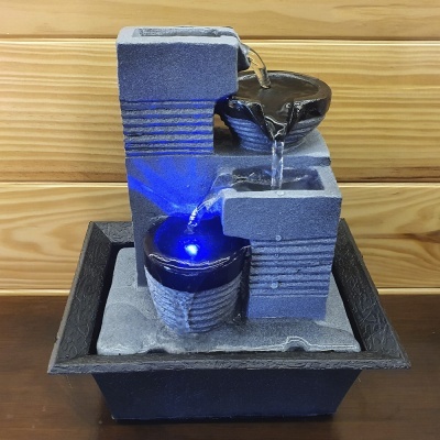 Fonte decorativa cinzenta com água e luz azul em base preta sobre mesa de madeira.