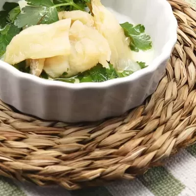 Tigela branca com peixe cozido e folhas verdes sobre suporte de palha trançada.