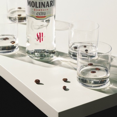Garrafa Molinari Sambuca Extra com copos de água e grãos de café em superfície branca
