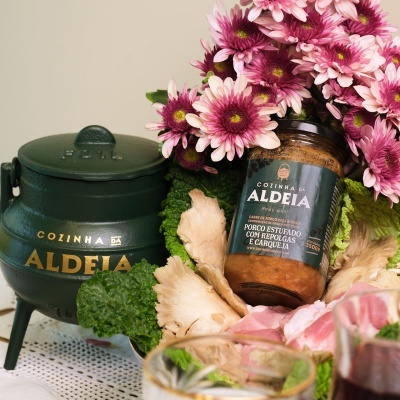 Frasco de comida tradicional em vidro com rótulo preto e dourado, vaso verde metálico, flores cor-de-rosa e vinho tinto sobre toalha branca