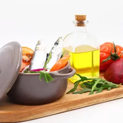 Panela com peixes e vegetais ao lado de garrafa de azeite e legumes em tabuleiro de madeira