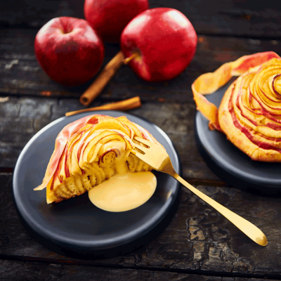 Dois pastéis de maçã em pratos pretos com maçãs vermelhas e paus de canela ao fundo
