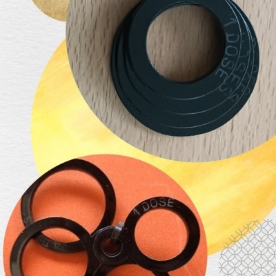 Peças plásticas pretas em anéis interligados com texto '1 DOSE' sobre fundo de madeira e papel colorido.