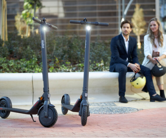 Segway-Ninebot E22 Dois trotinetes elétricos negros com luzes acesas e a marca ninebot, com duas pessoas sentadas ao fundo a segurar capacetes numa área urbana
