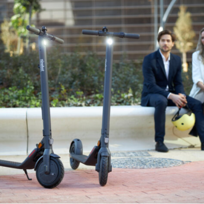 Dois trotinetes elétricos negros com luzes acesas e a marca ninebot, com duas pessoas sentadas ao fundo a segurar capacetes numa área urbana