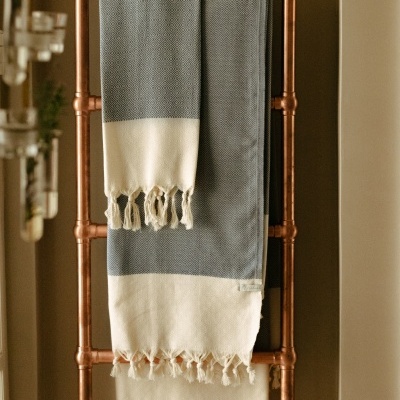 Manta cinza e branca com franjas pendurada em estrutura metálica de cor cobre