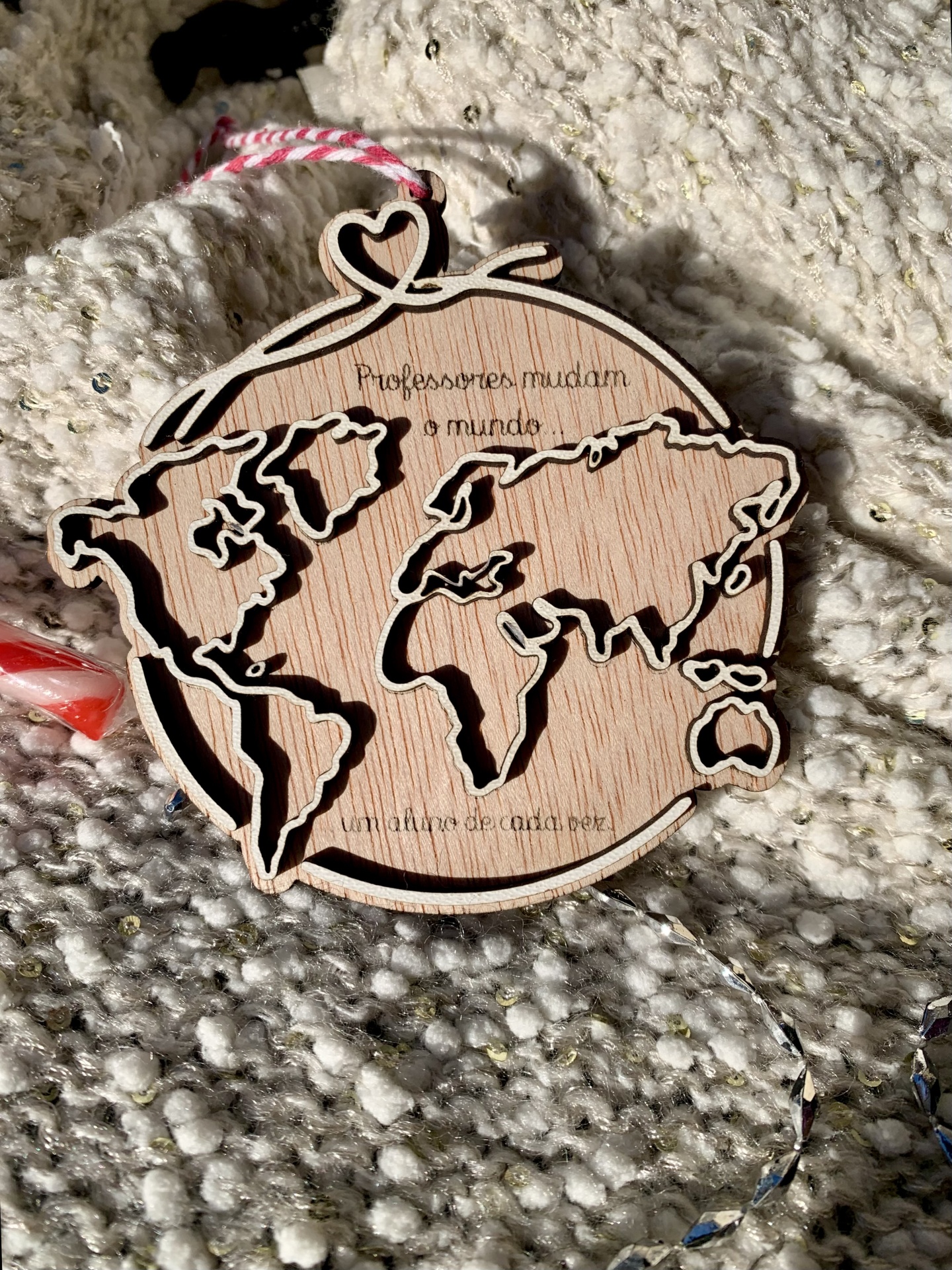 Ornamento redondo em madeira com mapa mundi recortado e texto gravado, pendurado sobre tecido cinzento com brilhos