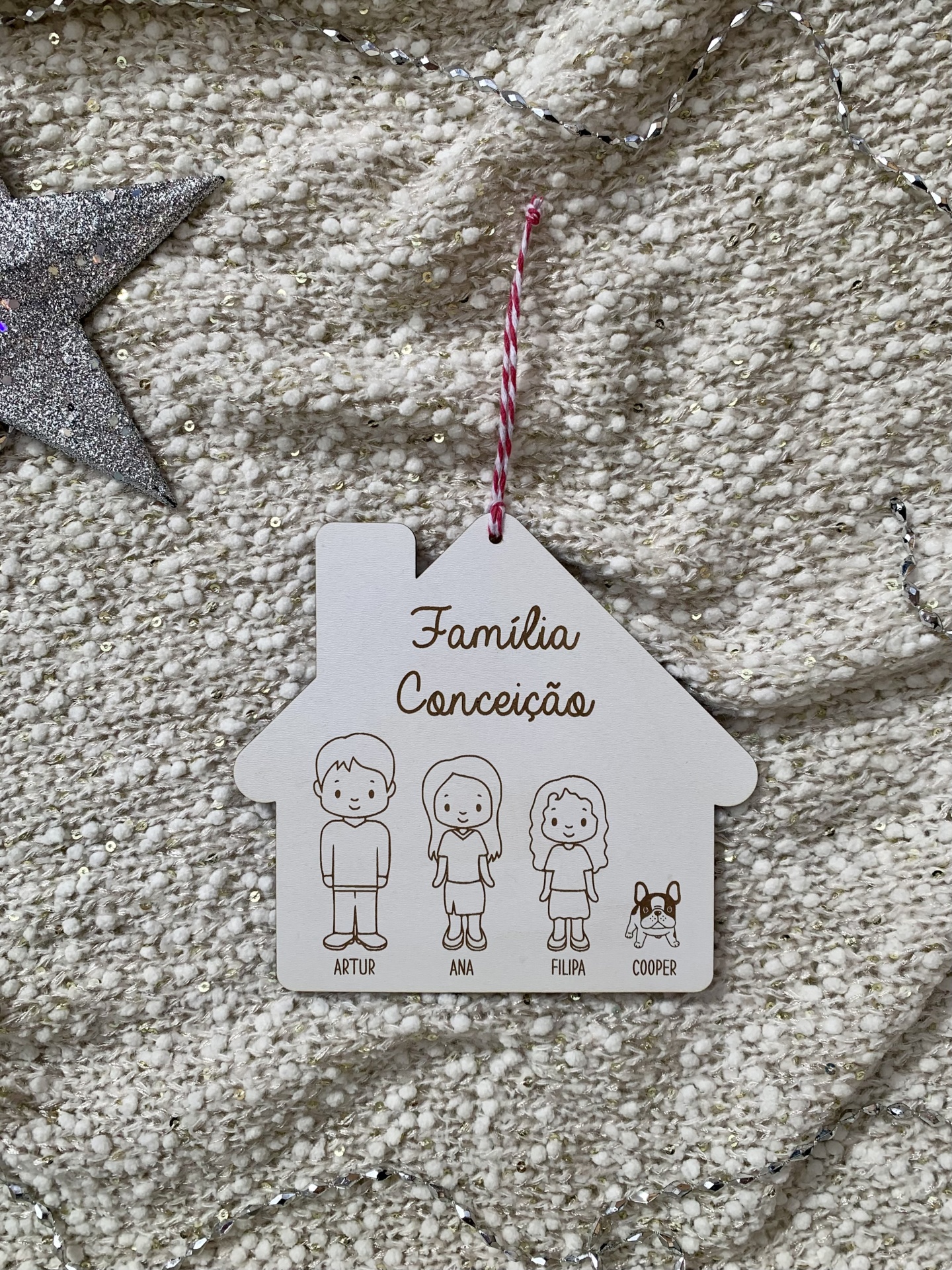 Placa decorativa branca em forma de casa com desenhos de pessoas e cão e nomes pendurada em tecido bege