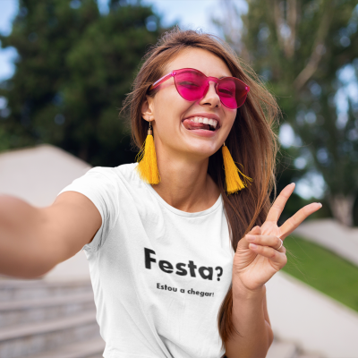 Mulher a tirar selfie com camiseta branca escrita 'Festa? Estou a chegar!' e acessórios coloridos.