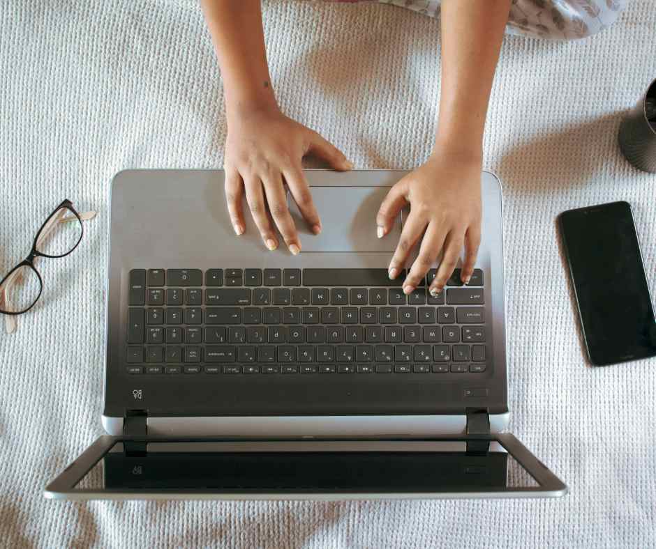 Computador portátil cinza com teclado preto, mãos digitando, óculos e smartphone ao lado