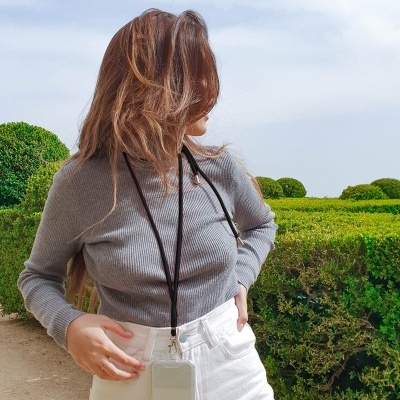 Pessoa com camisola cinzenta e calças brancas, bolsa transparente pendurada no pescoço, arbustos ao fundo