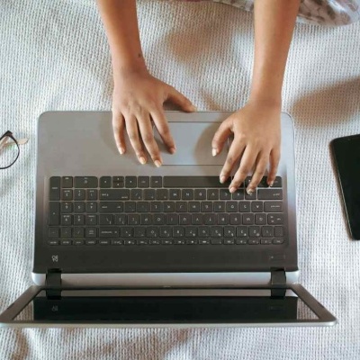 Computador portátil cinza com teclado preto, mãos digitando, óculos e smartphone ao lado