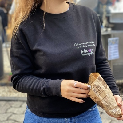 Mulher a usar sweatshirt preta com texto bordado e calças de ganga, segurando saco de papel