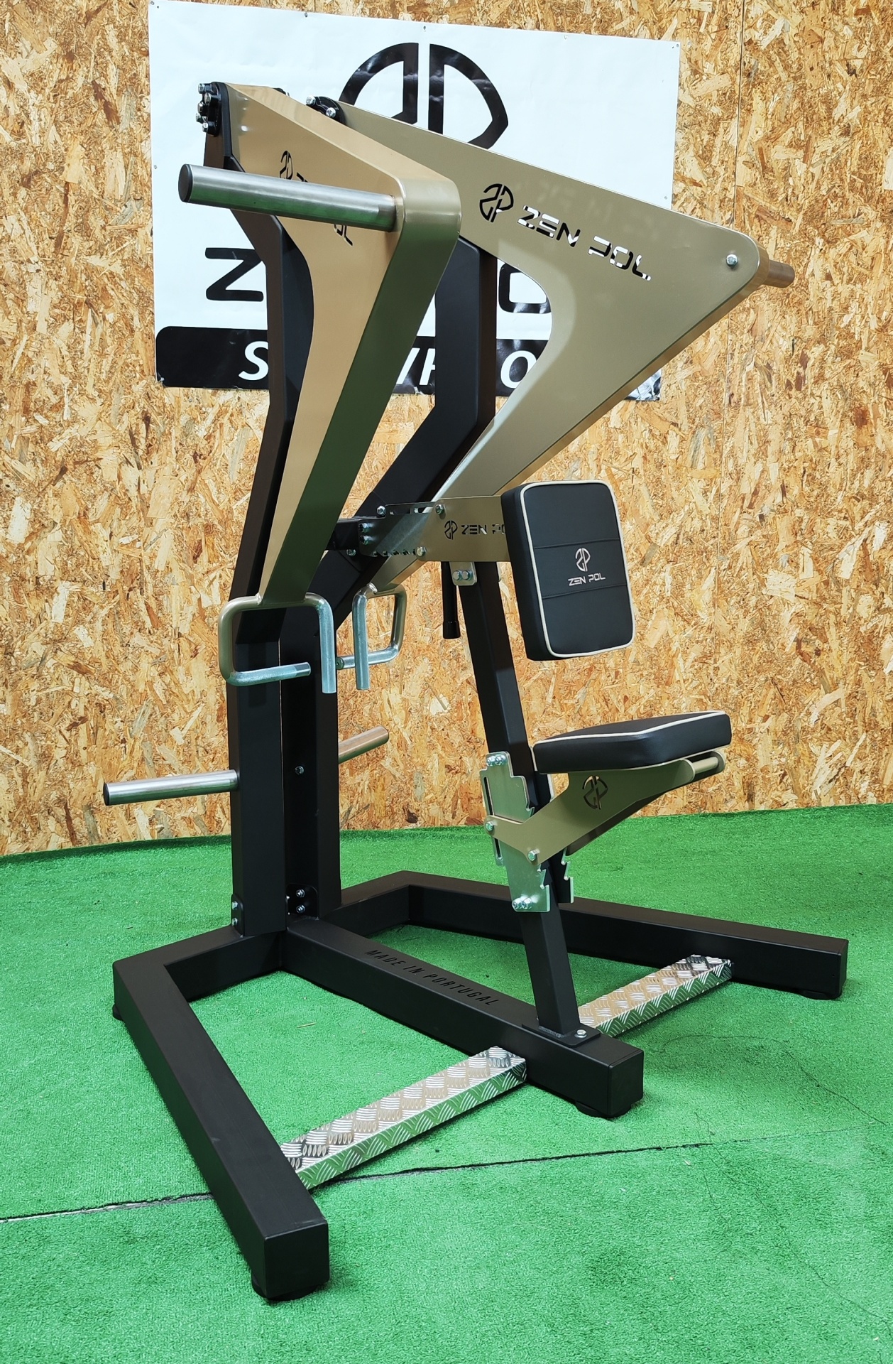 Equipamento de musculação metálico preto e dourado com assento acolchoado em fundo verde e madeira