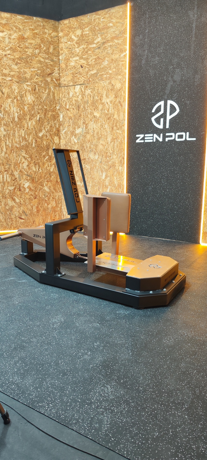 Máquina de ginásio bege e preta com logo ZEN POL num espaço com paredes de madeira OSB e parede preta.