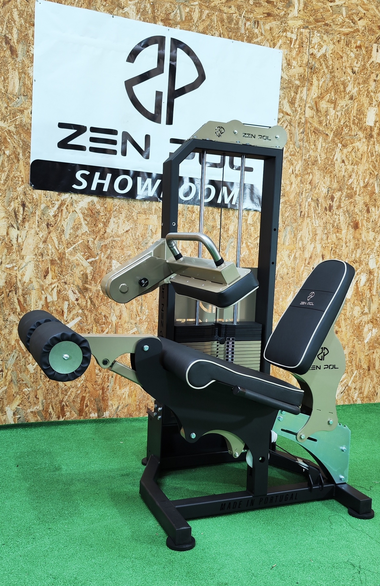 Máquina de exercício de pernas em metal preto e bege, estofos pretos com logotipo ZEN POL, sobre tapete verde com parede de madeira e cartaz ZEN POL SHOWROOM