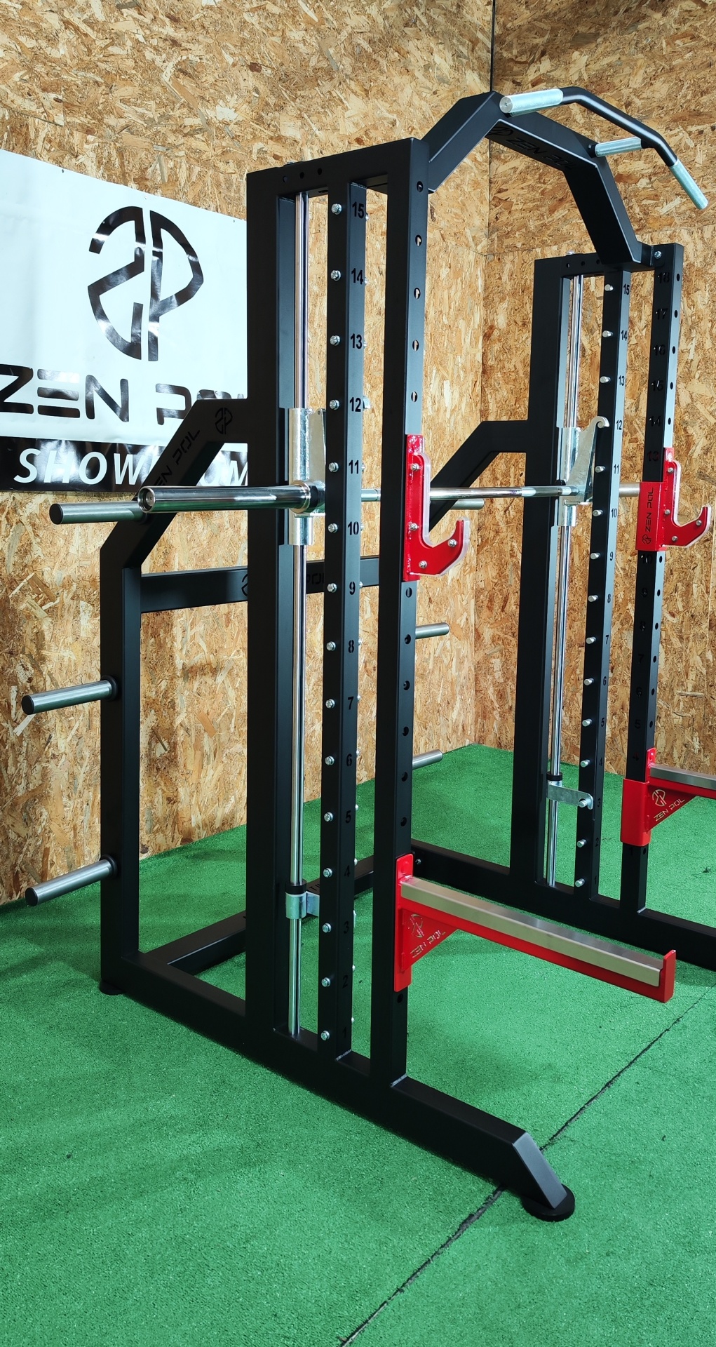 Estrutura metálica de musculação preta com detalhes vermelhos, barras prateadas, em ambiente com chão verde e parede de madeira