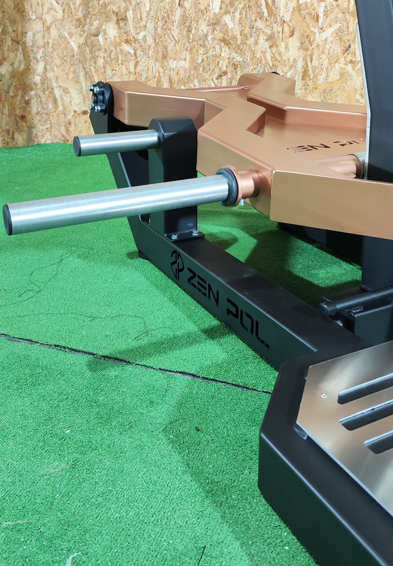 Equipamento de ginásio preto e cobre com marca Zen Pol sobre tapete verde e fundo de madeira