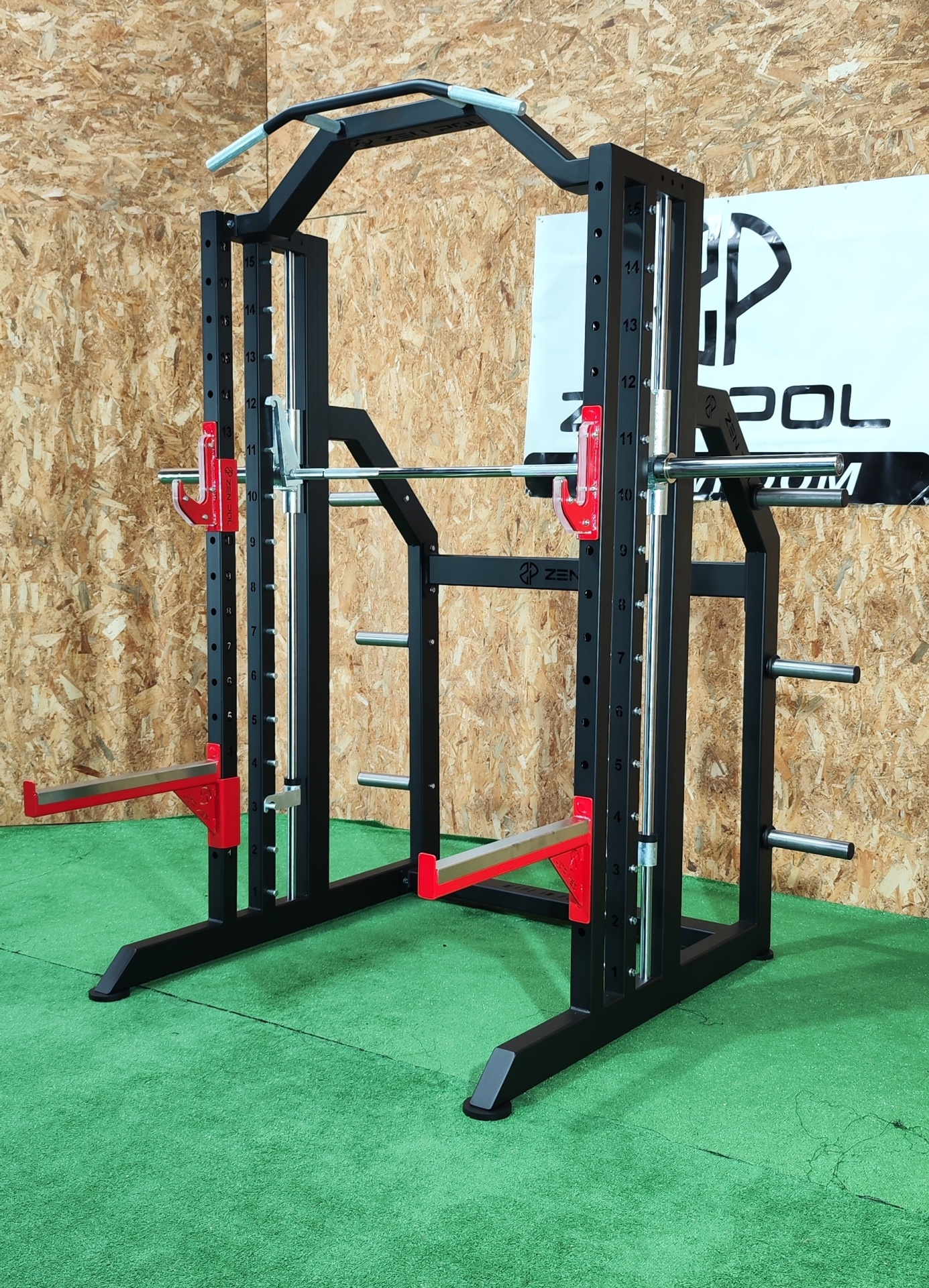 Máquina Smith machine para exercícios com estrutura metálica preta e detalhes vermelhos, chão verde e parede de madeira