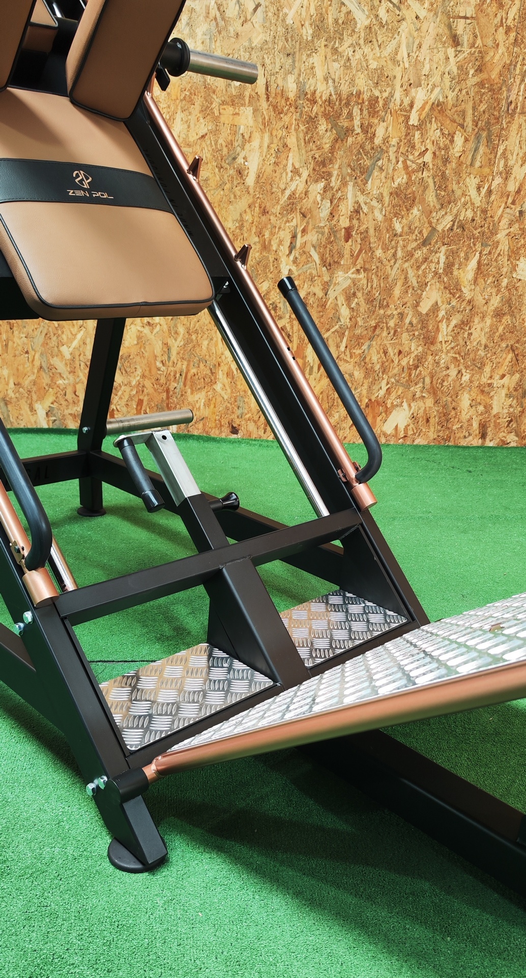 Máquina de leg press com estrutura preta e cobre num piso de grama artificial verde
