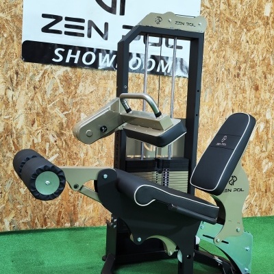 Máquina de exercício de pernas em metal preto e bege, estofos pretos com logotipo ZEN POL, sobre tapete verde com parede de madeira e cartaz ZEN POL SHOWROOM